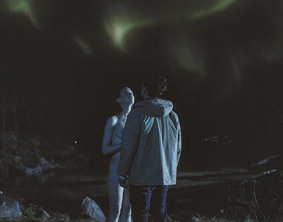 Naked woman and man in anorak staring up at aurora borealis