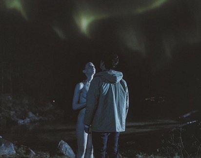 Naked woman and man in anorak staring up at aurora borealis