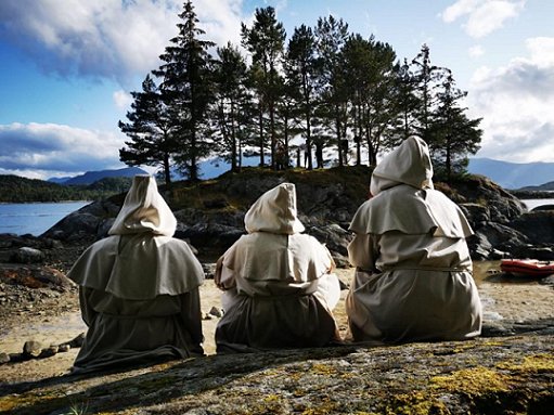 Three hooded figures sitting facing a wooded copse in background