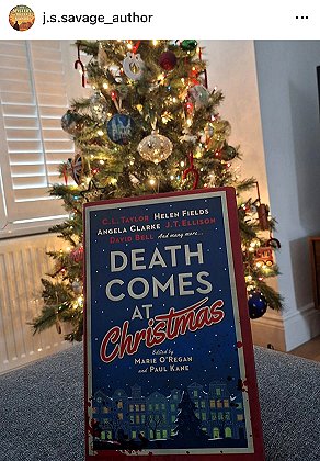 screenshot from @j.s.savage_author. Image shows a copy of Death Comes at Christmas, edited by Marie O'Regan and Paul Kane, standing up against a decorated mini Christmas tree on a dark grey surface.