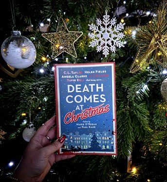 photograph showing a woman's hand holding a copy of Death Comes at Christmas, edited by Marie O'Regan and Paul Kane, up against a decorated Christmas tree