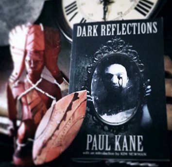 photograph of a copy of Dark Reflections by Paul Kane, propped against a statue of a spear-wielding warror wearing an ornate headdress