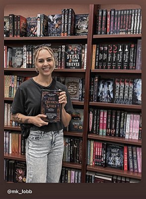 photograph of a smiling MK LObb standing in a bookstore, holding up a copy of These Dreaming Spires, edited by Marie O'Regan and Paul Kane