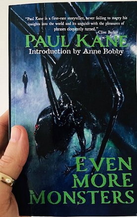 photograph of a man's hand holding up a copy of Even More Monsters by Paul Kane, introduction by Anne Bobby. Book cover shows a black spider-like monster in the foreground, with a man in a black suit standing in the background against a blue background