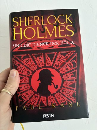 photograph showing a man's hand holding a copy of the German edition of Sherlock Holmes and the Servants of Hell by Paul Kane up against a white background