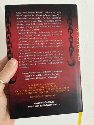photograph of a man's hand holding a copy of the German edition of Sherlock Holmes and the Servants of Hell by Paul Kane up against a white background, showing the back cover of the book