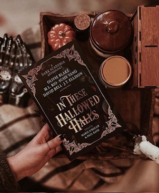 image of a woman's hand holding a copy of In These Hallowed Halls, edited by Marie O'Regan and Paul Kane, over a box containing a pumpkin ornament, a brown jar a cork-lidded jar and a metal lid. The box is lying on a cream and brown striped cloth, with a black hand ornament with white eyes on the palms