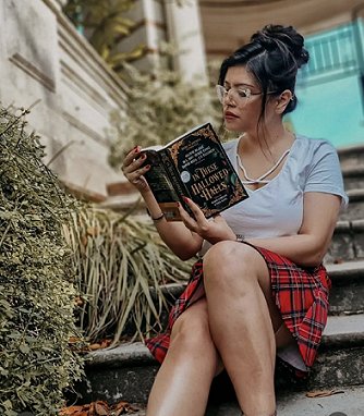 A photograph of a woman wearing a grey t-shirt and red plaid skirt,with glasses and with her dark hair in a bun, sitting on steps reading a copy of In These Hallowed Halls, edited by Marie O'Regan and Paul Kane
