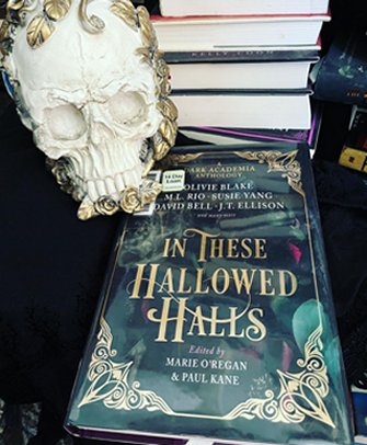 image shows a copy of In These Hallowed Halls, edited by Marie O'Regan and Paul Kane, lying on a black surface. A skull ornament is to its left