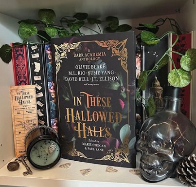 image shows a copy of In These Hallowed Halls, edited by Marie O'Regan and Paul Kane, standing in front of a row of books. An In These Hallowed Halls bookmarks stands to the left, and the shelf also holds a key, small heart-shaped bits of paper with text on, and a glass skull jar and pine cone. A plant grows along the top of the shelf at the back