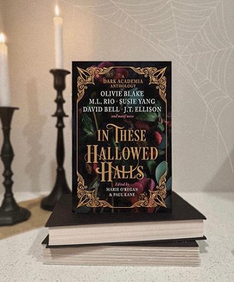 A copy of In These Hallowed Halls standing on two flat books, on a cream table in front of two ornate candlesticks with lit white candles
