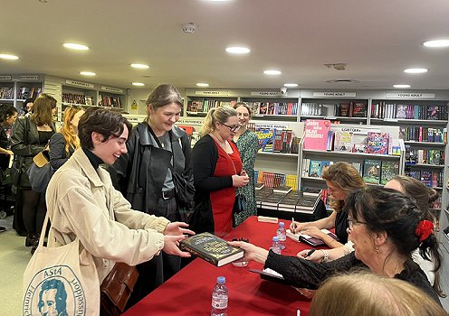 Photo of the queue to get copies of In These Hallowed Halls signed. Helen Grant is handing a signed book back to a customer. 