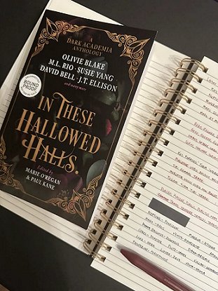 photograph of a proof copy of In These Hallowed Halls, edited by Marie O'Regan and Paul Kane, lying on top of an open spiral bound notebooks filled with story notes, and a dark red pen lying on top of the notebook