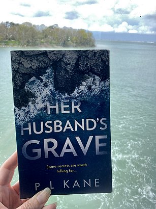 a copy of the book Her Husband's Grave by PL Kane, being held up against a backdrop of the sea with wooded coast behind