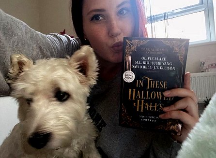 Image of a woman sitting down with a small white dog beside her. She's holding a copy of In These Hallowed Halls, edited by Marie O'Regan and Paul Kane, 