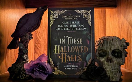 image showing a copy of In These Hallowed Halls, edited by Marie O'Regan and Paul Kane, standing in front of a wooden background, between a wooden skull on the right and a statue of a crow standing on a branch on the left, alongside a purple flower. 