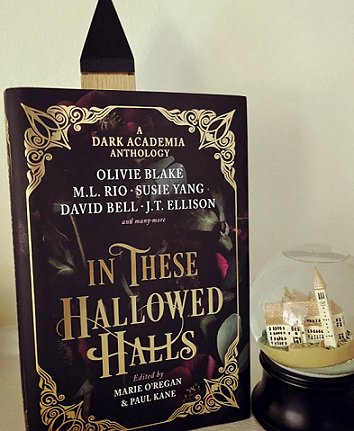 photograph of a standing copy of In These Hallowed Halls, edited by Marie O'Regan and Paul Kane, against a cream background. A castle ornament in a glass dome is alongside