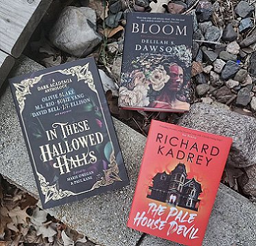 display of three books lying on a plank on the ground. Books are: In These Hallowed Halls, edited by Marie O'Regan and Paul Kane, The Pale House Devil by Richard Kadrey and Bloom by Delilah Dawson