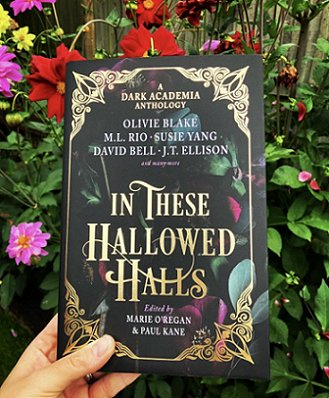 photograph showing a hand holding a copy of In These Hallowed Halls, edited by Marie O'Regan and Paul Kane, up against a background of red, pink and yellow flowering plants