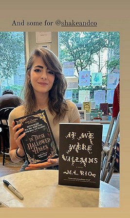 Image of ML Rio sitting behind a standing copy of If We Were Villains while holding a copy of In These Hallowed Halls, edited by Marie O'Regan and Paul Kane