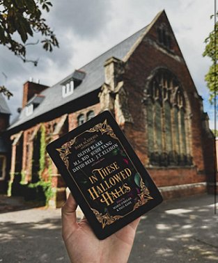 A hand holding a copy of In These Hallowed Halls, edited by Marie O'Regan and Paul Kane, in front of an old church building