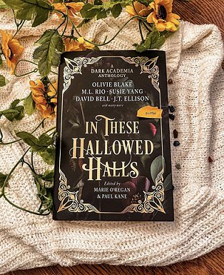 photograph of a copy of In These Hallowed Halls, edited by Marie O'Regan and Paul Kane, lying on a cream knitted cloth on a wooden background, with yellow flowers and green leaves underneath