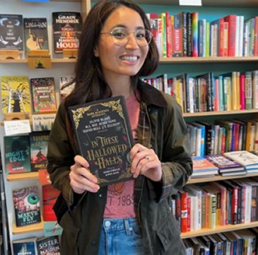 photograph of author Olivie Blake standing in front of bookshelves, holding up a copy of In These Hallowed Halls, edited by Marie O'Regan and Paul Kane