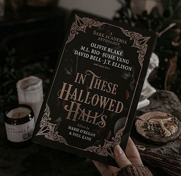 image of a woman's hand holding up a copy of In These Hallowed Halls, edited by Marie O'Regan and Paul Kane. Behind the book are candles and coaster on a brown cloth
