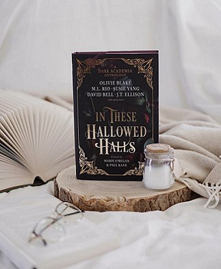 photograph of a copy of In These Hallowed Halls, edited by Marie O'Regan and Paul Kane, standing on a wooden base, in front of an open book and beside a glass jar of milk, on a beige knitted throw. In front, on a white cloth, is a pair of glasses.