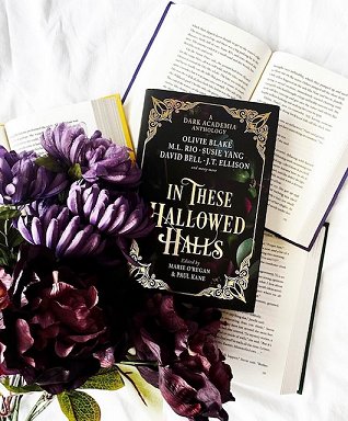 image shows a copy of In These Hallowed Halls, edited by Marie O'Regan and Paul Kane, lying on top of three open books on a white background, with dark red and purple flowers beside it