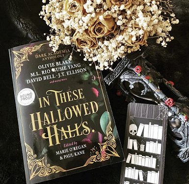 A proof copy of In These Hallowed Halls, edited by Marie O'Regan and Paul Kane, lying on a black cloth, beside a black bookmark decorated with a cartoon of a skull and books on shelves. ALso beside an ornate black key decorated with red roses, and a posy of cream flowers - roses and gypsophila