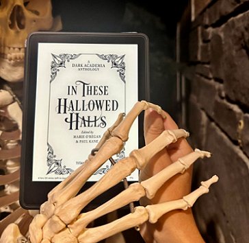 image of a hand holding a tablet device, showing the title page of In These Hallowed Halls, edited by Marie O'Regan and Paul Kane. A skeleton is holding its hands over the hand holding the tablet