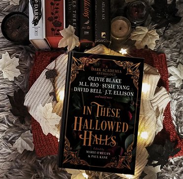 A copy of In These Hallowed Halls, edited by Marie O'Regan and Paul Kane, against a cream and red knitted cloth, decorated with creaml eaves and a brass key, with books in the background