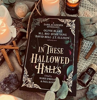 image shows a copy of In These Hallowed Halls, edited by Marie O'Regan and Paul Kane, lying on a pale green knitted cloth, surrounded by cinnamon sticks, lit candles and an amethyst geode. A small brown glass bottle with a metal lid lies to the right, and there are leaves around the edge of the display