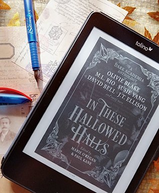 image shows a black tablet on a table littered with postcards and a blue fountain pen with a blue and red lid beside it, showing the black and white cover of In These Hallowed Halls, edited by Marie O'Regan and Paul Kane