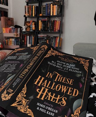 An open copy of In These Hallowed Halls, edited by Marie O'Regan and Paul Kane, lying to show the cover, against a backdrop of bookshelves