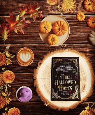 Display featuring a copy of In These Hallowed Halls, edited by Marie O'Regan and Paul Kane, lying on a log on a wooden table, alongside orange flowers, a purple candle, a hot drink with a heart design on top, three little pumpkins on a cream plate, and autumn leaves