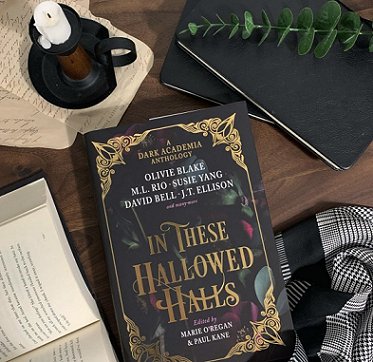 image shows a smiling woman with long brown hair, dressed in a black slip and brown cardigan, holding up an open copy of In These Hallowed Halls, edited by Marie O'Regan and Paul Kane