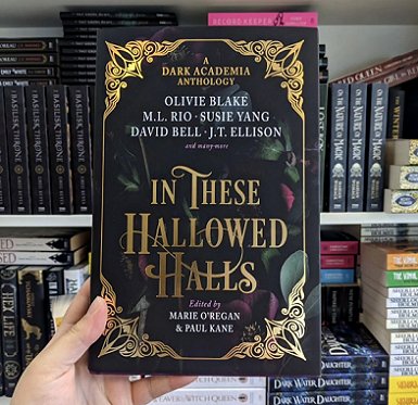Picture of a hand holding a copy of In These Hallowed Halls, edited by Marie O'Regan and Paul Kane, against a background of filled bookshelves in the Titan offices