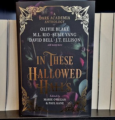 photograph of a copy of In These Hallowed Halls, edited by Marie O'Regan and Paul Kane, standing in front of a row of books, page edges facing out