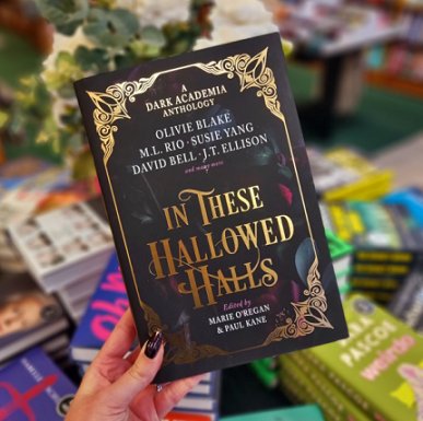 A woman's hand with black nail varnish, holding up a copy of In These Hallowed Halls, edited by Marie O'Regan and Paul Kane, against a background of a display of other books and a floral centrepiece