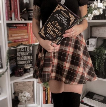 photograph of a woman in a short plaid skirt and black top, stockings, holding a copy of In These Hallowed Halls, edited by Marie O'Regan and Paul Kane, whilst standing in front of bookshelves decorated with books and flowers