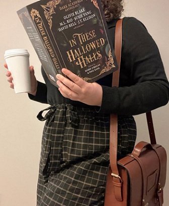 image shows a woman in a black top and black and white check trousers holding a white styrofoam cup, with a brown handbag over her left shoulder. In her left hand, she's holding up a copy of In These Hallowed Halls, edited by Marie O'Regan and Paul Kane