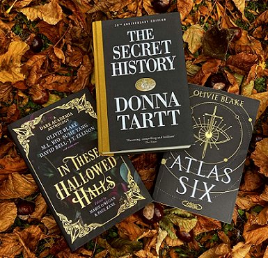 Three books lying on a bed of autumn leaves. The books are: Atlas Six by Olivie Blake, The Secret History by Donna Tartt and In These Hallowed Halls, edited by Marie O'Regan and Paul Kane
