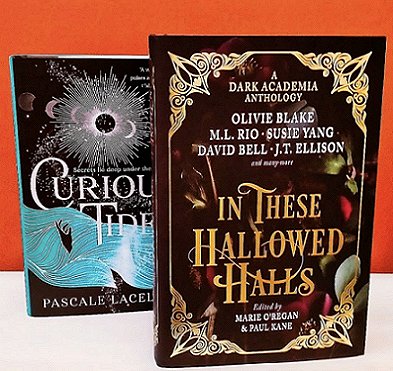 two books standing on a white surface against an orange background. The books are Curious Tides by Pascale Lacelle and In These Hallowed Halls, edited by Marie O'Regan and Paul Kane