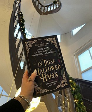 image of a woman's hand holding a copy of In These Hallowed Halls, edited by Marie O'Regan and Paul Kane, up against a black spiral staircase in a white painted hall