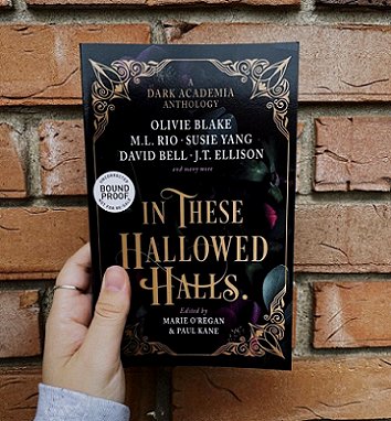 A woman's hand holding a copy of In These Hallowed Halls, edited by Marie O'Regan and Paul Kane, up against a brick wall