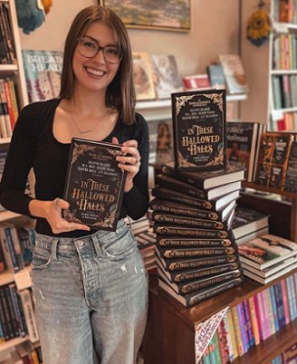 photograph of Kelly Andrew in a bookshop beside stacked copies of In These Hallowed Halls, edited by Marie O'Regan and Paul Kane. She's wearing a black long sleeved top and pale blue jeans, and holding up a copy of In These Hallowed Halls