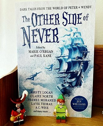 photograph of a standing copy of the book The Other Side of Never, edited by Marie O'Regan and Paul Kane, on a wooden surface. In front are Lego figures of Captain Hook and Peter Pan