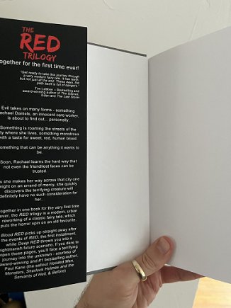 image of a man's hand holding open a copy of The RED Trilogy by Paul Kane, to show the inside flap text.